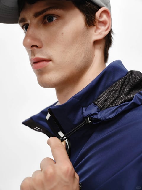 The image is a photorealistic close-up portrait of a young man with a serious, focused expression, framed from the mid-chest up against a plain, bright white background. The composition is angled, with the subject looking off-camera to the left, his face in three-quarter profile. He wears a dark grey baseball cap partially visible at the top edge and a deep navy blue technical jacket. His right hand is raised, fingers gently gripping the black zipper pull at the jacket's collar. A major visual element is the contrast between the jacket's smooth, matte navy exterior and the intricate, dark mesh lining with a fine, white dotted pattern visible inside the collar. The lighting is soft and directional, coming from the upper left, creating gentle highlights on his cheekbone, nose, and the jacket's shoulder, with subtle, natural shadows under his jaw and in the fabric folds, conveying a clean, studio-shot mood. The color palette is minimalist and cool, dominated by the deep navy blue of the jacket, the neutral skin tones, and the bright white background, with high contrast between the dark fabrics and the light setting. The texture rendering is highly detailed, emphasizing the smooth, water-resistant look of the jacket's outer shell, the soft, slightly porous texture of the skin, and the precise, grid-like pattern of the inner mesh lining. There is no obvious readable text in the image. The overall mood is one of modern, technical precision and quiet intensity, suitable for a style reference focusing on contemporary menswear or technical apparel details.