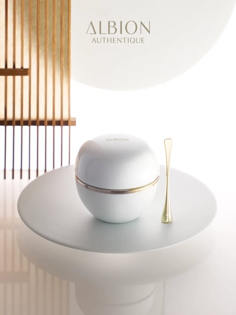 This image features a photorealistic, elegantly composed product shot of a luxury skincare set, centered on a smooth, off-white circular pedestal. The main subject is a spherical, glossy white jar with a subtle metallic gold band encircling its middle, topped with a domed lid embossed with the word 'ALBION'. To its right stands a slender, elongated gold spatula. The scene is set against a minimalist, bright white background with a soft, circular gradient behind the product, enhancing its prominence. To the left, a vertical architectural element of thin, warm-toned wooden slats creates a subtle Japanese-inspired frame, adding depth and a natural texture contrast. Visible text includes the brand name 'ALBION' in a refined, uppercase sans-serif font at the top center of the image, with the word 'AUTHENTIQUE' in smaller uppercase letters directly below it, both in a soft, muted gold color. The lighting is soft and diffused, appearing to come from the upper left, casting gentle, elongated shadows that emphasize the jar's smooth curvature and the spatula's polished finish, creating a serene, luxurious, and clean atmosphere. The color palette is restrained and sophisticated, dominated by pure whites, creamy off-whites, and warm metallic golds, with high contrast between the bright whites and the deep, soft shadows, resulting in a cool-to-neutral color temperature. Textures are meticulously rendered: the jar has a flawless, high-gloss ceramic-like surface, the pedestal is a matte, smooth stone or composite, the spatula is a polished, reflective metal, and the wooden slats have a fine, linear grain. The composition is balanced and minimalist, using a shallow depth of field to keep the product in sharp focus while softly blurring the background elements, with the circular pedestal and jar creating a strong central focal point against the linear verticality of the wooden screen.
