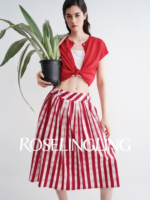 The image is a full-body, studio-shot fashion photograph featuring a young woman standing against a plain, bright white background, creating a clean and minimalist composition focused entirely on the subject. The overall visual aesthetic is photorealistic and crisp, with a high-contrast, vibrant color palette dominated by bold red and crisp white, accented by the deep green of a potted plant she holds in her right hand. The lighting is soft and directional, appearing to come from the front-left, casting gentle shadows that define her form and the textures of her clothing without harshness. Her outfit consists of a red, short-sleeved, button-up cardigan knotted at the waist over a white bandeau top, paired with a high-waisted, A-line midi skirt featuring wide, vertical red and white stripes and a diagonal-striped waistband. The textures are distinct: the soft knit of the cardigan, the smooth cotton of the top, and the crisp, structured fabric of the pleated skirt. The composition is centered and balanced, with the subject's upright posture and the vertical lines of the skirt creating strong vertical movement, while the plant adds an organic, asymmetrical element. The text 'ROSELINGLING' is superimposed in a large, elegant, white serif font across the lower third of the image, overlapping the skirt; it serves as a brand identifier or title within the fashion editorial context. The mood is modern, fresh, and stylish, with a clean, high-energy feel conveyed through the bright lighting and saturated primary colors.