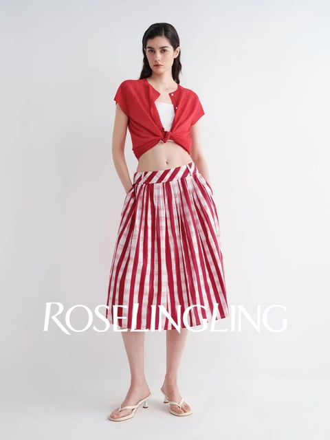 The image presents a full-body studio photograph of a young woman with long dark hair, standing against a seamless white background. She wears a bright red short-sleeved shirt, knotted at the waist over a white top, exposing her midriff, paired with a high-waisted, midi-length skirt featuring bold red and white vertical stripes on a pleated gingham fabric. The composition is centered and symmetrical, with a straight-on camera angle capturing the entire figure from head to toe. The lighting is bright, even, and diffuse, characteristic of high-key studio lighting, creating soft shadows and a clean, commercial aesthetic. The color palette is dominated by vibrant red and crisp white, with high saturation and strong contrast between the stripes and the neutral background. The visual style is photorealistic with a focus on fashion styling, emphasizing the texture of the crisp cotton shirt and the structured, pleated drape of the gingham skirt. The mood is fresh, summery, and playful, with a clean, energetic vibe. The word 'ROSELINGLING' is prominently displayed in a large, white, serif typeface across the lower portion of the image, overlaying the skirt, functioning as a brand watermark or logo.