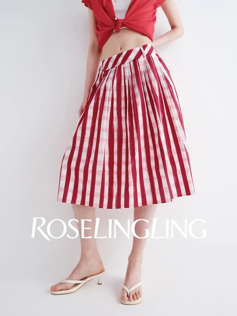 The image is a vertically framed, photorealistic studio-style photograph focusing on the lower torso and legs of a person against a plain, seamless white background. The main subject is wearing a vibrant red and white vertically striped, pleated midi skirt, with a tied red short-sleeved cardigan and white top partially visible at the waist. The composition is clean and minimalist, with the subject centered, creating a clear focus on the garment's form and pattern. The color palette is dominated by a high-contrast, saturated pairing of bright candy-apple red and crisp white, with the lighting appearing soft, diffused, and frontal, minimizing harsh shadows and evenly illuminating the textures. The skirt's material appears to be a lightweight, possibly cotton or linen blend, with a crisp hand feel evident in the sharp, vertical pleats that catch the light, creating subtle shadows within the folds and a sense of structured movement. The shape language is defined by the strong, repetitive vertical lines of the stripes and pleats, contrasting with the organic curves of the body and the diagonal lines created by the V-shaped waistband and the tied knot of the cardigan. The mood is playful, summery, and casually chic, with a fresh and energetic visual weight anchored by the bold pattern. Overlaid at the bottom center is the text 'ROSELINGLING' in a clean, elegant, white serif font, which serves as a brand watermark, identifying the likely source or designer of the clothing.