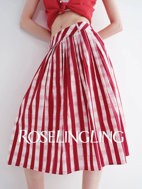 The image is a photorealistic studio-style shot focusing on a woman's lower torso and legs, showcasing a red and white vertically striped pleated skirt that falls to just above the knees, paired with a cropped red knitted top tied at the waist; the composition is tightly framed against a plain white background, emphasizing the garment's texture and form. Across the lower half of the skirt, the text 'ROSELINGLING' is prominently displayed in a bold, white, sans-serif font, serving as a brand identifier or watermark. The lighting is soft and diffuse, coming from the front-left, creating gentle highlights on the pleats and subtle shadows that enhance the skirt's volume and the fabric's slight sheen, suggesting a smooth, lightweight material. The color palette is dominated by vibrant, saturated reds and crisp whites with high contrast, evoking a fresh, energetic, and summery mood, while the composition uses a shallow depth of field to keep the skirt in sharp focus against the neutral background.