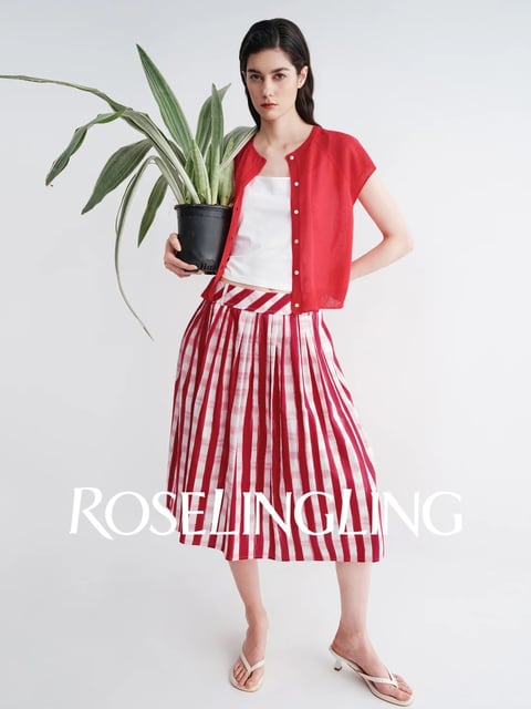 A full-length studio portrait captures a young woman standing against a plain, light gray background, presenting a casual, modern fashion look. The composition is centered and clean, with the subject holding a potted plant, creating a balanced and slightly playful arrangement. The image is photorealistic, rendered with sharp detail and a focus on texture and form. The color language is dominated by a vibrant, saturated red in the short-sleeved cardigan and the bold vertical stripes of the midi skirt, contrasted against the crisp white of the cropped tank top and the skirt's lighter stripes. The lighting is soft and diffused, coming from the front and slightly to the side, creating gentle shadows that define the subject's features and the folds of the clothing without harshness, contributing to a bright, airy mood. Texture is rendered with precision, from the subtle knit of the red cardigan and the smooth cotton of the tank top to the crisp, pleated fabric of the striped skirt and the organic, waxy leaves of the variegated plant in its simple black pot. The composition uses a straightforward, frontal framing with the subject's body creating a gentle S-curve, and the plant adds an organic, asymmetrical element. Shape language is a mix of the structured, geometric lines of the skirt's stripes and the cardigan's placket against the softer, organic curves of the subject's figure and the plant's leaves. The overall mood is fresh, confident, and casually chic, with a light, energetic atmosphere. A prominent text overlay in a bold, white, serif font reads 'ROSELINGLING', positioned across the lower third of the image, serving as a brand or editorial identifier and integrating graphically with the striped pattern of the skirt.