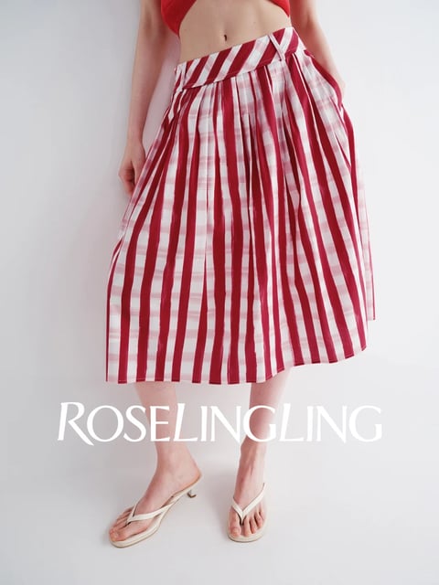 The image features a mid-length, pleated skirt as the central subject, photographed from the waist down on a model against a plain, light grey background. The composition is a clean, centered product shot focusing on the garment's fit and drape. The skirt is made of a crisp, likely cotton or linen-blend fabric, with a striking pattern of bold, vertical red stripes alternating with wider panels of a subtle, tonal plaid in shades of red, pink, and white. The waistband is wide and features a folded, asymmetrical detail with diagonal stripes that contrast with the vertical lines of the skirt body. The model wears simple, cream-colored, low-heeled thong sandals. A prominent text overlay reads 'ROSELINGLING' in a white, elegant serif font, positioned across the lower third of the image, serving as a brand identifier. The visual aesthetic is clean, modern, and product-focused, with a photorealistic rendering that highlights the fabric's texture and the garment's structured yet flowing silhouette. The color language is dominated by a vibrant, warm red against neutral whites and greys, creating high contrast and a fresh, summery feel. The lighting is bright, soft, and diffuse, likely from a large frontal source, minimizing harsh shadows and evenly illuminating the fabric's details and colors. The texture of the skirt appears smooth and structured, with sharp, pressed pleats that create rhythmic, geometric lines. The overall mood is chic, effortless, and contemporary, emphasizing the garment's bold pattern and tailored design within a minimalist setting.