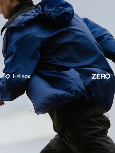 This image features a dynamic, low-angle, photorealistic shot of a person in motion, seen from the back and slightly below, emphasizing the bulk and texture of their outerwear. The dominant subject is a person wearing a voluminous, dark navy blue hooded jacket and black technical pants, set against a plain, light grey background that suggests an overcast sky. The composition is tightly cropped, focusing on the torso and upper legs, with the subject angled diagonally, creating a sense of forward momentum. The visual aesthetic is clean, modern, and technical, with a focus on material details. The color palette is cool and restrained, dominated by deep navy blue and black, with high contrast against the neutral light grey backdrop; the lighting is soft and diffused, likely simulating overcast daylight, which evenly illuminates the jacket's surface, revealing its fine, slightly wrinkled, matte fabric texture without harsh shadows. Visible text appears as white, sans-serif logos overlaid on the jacket: the word 'Helinox' with a diamond icon is positioned on the left sleeve, and the word 'ZERO' is on the right side of the back panel, both playing a clear branding role. The shape language combines the organic, soft folds of the jacket's puffy insulation with the structured, geometric lines of the logos. The overall mood is one of minimalist, functional athleticism and technical performance.