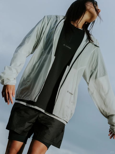 The image presents a low-angle, medium shot of a person outdoors against a clear, pale blue sky, capturing a dynamic, upward-looking composition that emphasizes movement and casual athletic style. The subject is wearing a layered outfit consisting of a semi-sheer, lightweight, pale grey or off-white windbreaker jacket, zipped open, over a black half-zip performance shirt and black athletic shorts with a subtle, crinkled nylon texture. The windbreaker's fabric is translucent, revealing the dark shirt underneath and showing fine wrinkles and folds that suggest a thin, technical material, while its sleeves are slightly bunched at the wrists. The black shirt features a small, white, geometric logo with the text 'Helinox' printed horizontally on the left chest, and the same 'Helinox' logo and text appear in black on the lower left panel of the jacket. The person's head is tilted back and to the right, with dark hair falling across the face, and their arms are extended outward to the sides, creating a sense of openness and energy. The lighting is bright, natural, and slightly harsh, coming from above and to the side, casting sharp shadows on the clothing folds and highlighting the translucent quality of the jacket fabric. The color palette is muted and cool, dominated by the pale blue sky, the off-white jacket, and the deep black of the inner layers, with a focus on matte and semi-transparent textures. The composition uses a low camera angle to create a sense of height and aspiration, with the subject's body forming diagonal lines that lead the eye upward, and the depth is shallow, keeping the focus sharply on the clothing details against the simple sky background.