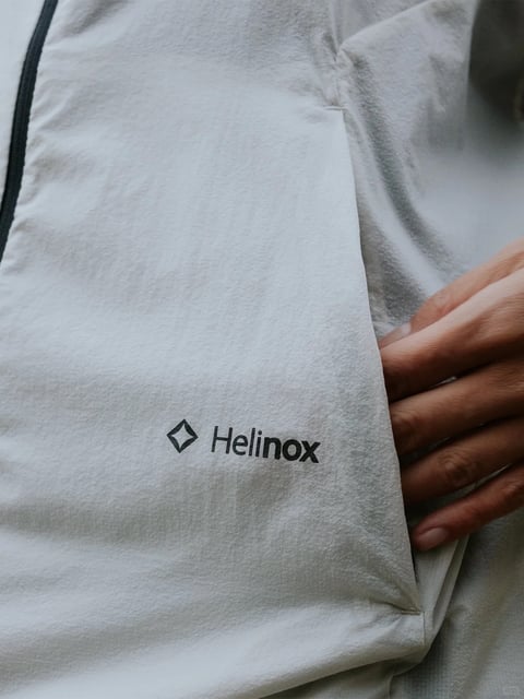 This image is a close-up, photorealistic shot focusing on the lower portion of a light gray, soft-textured garment, likely a fleece or pullover, featuring a prominent black Helinox logo. The composition is tightly framed, emphasizing the fabric's plush surface and the placement of the branding, with a person's hand resting gently on the garment's surface to the right, adding a sense of scale and interaction. The dominant color palette is muted and cool, centered around varying shades of light heather gray, with the crisp black logo providing a high-contrast focal point; the lighting is soft and diffused, appearing to come from above, which gently highlights the fabric's nap and creates subtle shadows in its folds, contributing to a calm, tactile atmosphere. The texture is rendered with meticulous detail, conveying a soft, fuzzy, and warm material feel typical of fleece, while the overall composition uses shallow depth of field to keep the logo and immediate fabric sharp, with the background and hand slightly softer, guiding the viewer's eye directly to the brand element.