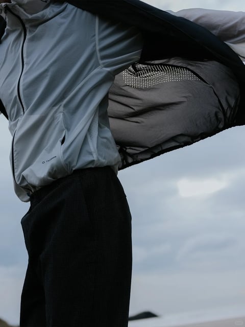 The image presents a mid-action, close-up shot of a person wearing technical outdoor apparel, with a dynamic composition that emphasizes movement and functionality. The subject is shown from the torso down to the upper thighs, captured in a low-angle perspective against a softly blurred, overcast sky background, suggesting an outdoor, possibly coastal or windy environment. The primary visual elements are the layered clothing: a light grey, long-sleeved technical base layer with subtle perforated venting details and a small 'Helinox' logo printed on the lower left side, and a dark, possibly black or deep charcoal, outer jacket that is billowing open dramatically to the right, revealing a dark interior lining with a distinctive pattern of small, bright white dots arranged in a grid. The person is also wearing dark, textured trousers with a fine, grid-like pattern. The overall visual aesthetic is photorealistic and cinematic, with a focus on material texture and motion. The color palette is muted and cool, dominated by shades of grey, black, and white, with low saturation and high contrast created by the bright white dot pattern against the dark jacket lining. The lighting is soft and diffused, characteristic of an overcast day, casting gentle shadows that define the folds and contours of the fabric without harsh highlights. Texture and material rendering are highly detailed, showcasing the smooth, synthetic feel of the base layer, the crisp, possibly waterproof shell of the jacket, and the nubby, woven texture of the trousers. The composition uses the diagonal sweep of the open jacket to create dynamic energy, framing the subject's torso while the background provides atmospheric depth. Shape language is a mix of the organic, flowing lines created by the wind-blown fabric and the geometric precision of the perforated dots and grid-patterned trousers. The mood is one of functional elegance and preparedness for the elements, conveying a sense of active movement and technical performance in a natural setting.
