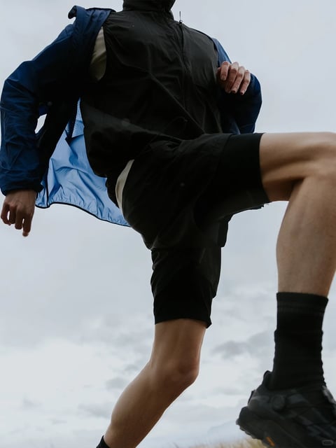 The image is a photorealistic, low-angle action shot capturing the lower torso and legs of a person in mid-stride, emphasizing dynamic movement. The composition focuses tightly on the subject's form against a bright, overcast sky, creating a strong sense of upward momentum and energy. The color palette is dominated by muted, cool tones: deep navy and black athletic wear contrast with the pale, diffuse gray-white of the sky, while the subject's skin tone adds a natural, warm mid-tone. The lighting is soft and diffuse, typical of an overcast day, providing even illumination without harsh shadows, which highlights the textures of the fabrics. The visual aesthetic is clean and modern, with a focus on functional athletic apparel. Textures are rendered with clear distinction: the windbreaker has a smooth, slightly glossy synthetic surface with visible seam lines and paneling, the shorts appear matte and flexible, and the socks and shoes show knit and rugged sole textures respectively. The composition uses a dynamic diagonal created by the subject's bent leg and angled torso, with the low perspective exaggerating the length and power of the movement. There is no readable text or logos visible on the clothing or in the background. The overall mood is one of active, focused energy and contemporary sportswear style, with a tactile emphasis on the performance materials of the outfit.