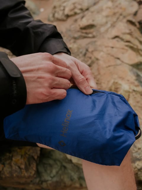 The image presents a close-up, slightly high-angle shot focused on a person’s hands interacting with a small, rectangular blue pouch set against a backdrop of rough, sun-baked rock surfaces. The composition is tight, framing the subject’s torso from the mid-chest down to the upper thigh, with the hands and pouch dominating the foreground and the textured rock forming a shallow depth of field in the background. The visual aesthetic is highly photorealistic, capturing the scene with naturalistic detail and rendering. The color language is dominated by the saturated, deep cobalt blue of the pouch, which contrasts sharply with the warm, earthy tones of the beige and tan rock formations and the muted black of the subject’s technical outerwear. Lighting appears soft and diffused, likely from an overcast sky or open shade, creating gentle shadows within the fabric folds and hand creases without harsh highlights, conveying a calm, outdoor ambiance. Texture and material feel are pronounced: the pouch exhibits a fine, matte ripstop nylon texture with subtle wrinkles, while the subject’s hands show detailed skin texture, knuckles, and veins. The rock background has a rough, granular, and uneven surface. Compositionally, the diagonal lines of the rock strata in the background lead the eye toward the central action of the hands opening the pouch, with the shallow depth of field isolating the subject. Shape language combines the soft, organic forms of the hands and fabric folds with the rigid, geometric rectangle of the pouch and the angular, fractured planes of the rock. The mood is one of focused, quiet preparation or rest during an outdoor activity, with a tactile, grounded atmosphere. Visible text includes the brand name 'Helinox' printed vertically in a muted, dark gray sans-serif font along the left side of the blue pouch, accompanied by a small, stylized logo to its left, identifying the product’s manufacturer.