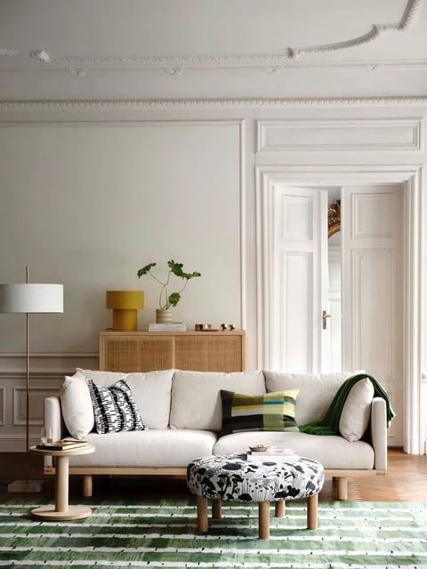 This is a photorealistic interior scene of a living room with a refined Scandinavian and modern Parisian apartment aesthetic, characterized by a calm, sophisticated atmosphere. The composition centers on a low-profile, light beige linen-upholstered sofa with a natural wood base, positioned on a green and white horizontally striped area rug. To the left of the sofa is a slender, round light-wood side table holding stacked books and a glass, while a cylindrical ottoman upholstered in a black-and-white botanical pattern sits directly in front. The background features a light sage-green wall with elegant classical white molding and paneling, including a tall white double doorway that opens into another brightly lit room with gilded mirror details. A natural wood credenza with woven cane doors stands against the back wall, topped with a mustard-yellow table lamp, a minimalist plant in a stone vessel, stacked books, and small brass objects. A tall, slim floor lamp with a white drum shade stands to the far left. The lighting is soft and diffuse, appearing to enter from the open doorway and unseen windows, casting gentle shadows and creating a serene, airy mood with a balanced color palette of muted greens, warm neutrals, and accents of ochre and brass. There is no obvious readable text visible in the image. The focus is sharp throughout, with clear rendering of textures including the linen sofa, woven cane, patterned rug, and smooth painted walls, emphasizing a tactile, livable yet curated space.