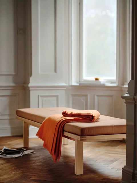 The image presents a serene, minimalist interior scene focused on a low, light wood bench with a beige cushion, positioned centrally against a backdrop of white, paneled walls with a tall window. A vibrant orange, textured throw blanket is casually draped over the bench, adding a pop of warm color. On the herringbone-patterned wooden floor, a few magazines are scattered near the bench's left leg. The visual aesthetic is photorealistic with a soft, painterly quality, emphasizing clean lines and subtle textures. The color language is dominated by a warm, neutral palette of whites, beiges, and natural wood tones, punctuated by the saturated orange of the blanket; the lighting is soft and diffused, likely natural light from the window, creating gentle shadows and a calm, inviting mood. The texture is rendered with tactile detail, from the woven fringe of the blanket to the smooth grain of the wood and the matte finish of the walls. Compositionally, the scene uses a balanced, slightly asymmetrical arrangement with strong vertical lines from the window and wall panels contrasting the horizontal bench, creating depth through layered planes. The shape language blends geometric elements (the rectangular bench, paneled walls) with organic touches (the draped blanket, scattered magazines). The overall mood is peaceful, quiet, and warmly minimalist, with a focus on tactile comfort and understated elegance. There is no obvious or readable text visible in the image.