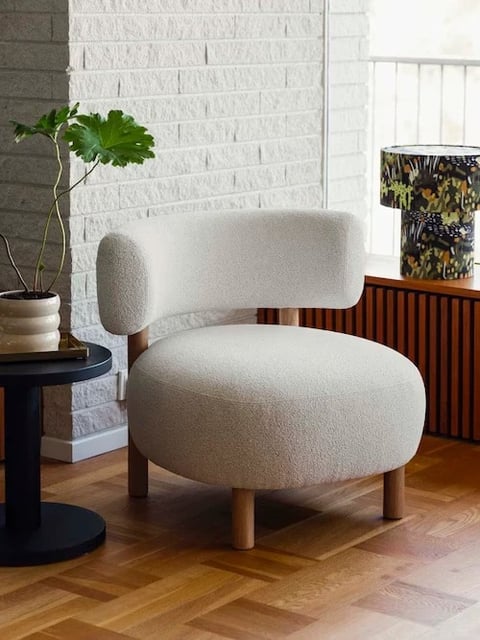 This photorealistic interior scene features a modern, curvaceous armchair as the central subject, positioned slightly off-center on a light brown herringbone-patterned wooden floor. The chair is upholstered in a soft, textured off-white bouclé fabric, with a rounded seat and a distinctive curved backrest supported by light wood legs, emphasizing organic, soft geometric shapes. To the left, a small, matte black circular side table holds a beige ceramic pot with a green leafy plant and a stack of books, while to the right, a fluted wooden cabinet sits beneath a window, topped with a cylindrical lamp featuring a dark, illustrated nature scene. The background includes a white-painted brick wall on the left and a bright window on the right, creating a balanced composition with clear spatial depth. The lighting is soft and diffused, appearing to come from the window on the right, casting gentle shadows and highlighting the tactile texture of the chair's fabric and the wood grain, contributing to a calm, inviting atmosphere with a neutral color palette of off-white, warm wood tones, black accents, and touches of green.