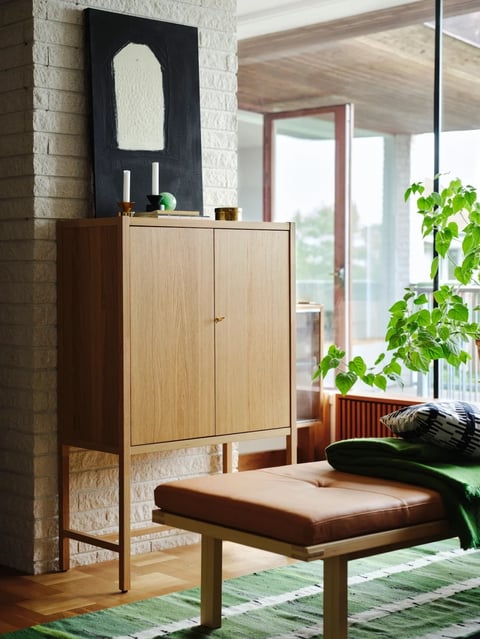 This photorealistic interior scene features a light oak, two-door cabinet with a slender, tapered frame and a single brass pull as the central subject, positioned against a textured, whitewashed brick pillar. The composition is a medium shot with a clear foreground (a brown leather bench and green rug), midground (the cabinet), and background (large windows and a glimpse of another room), creating a sense of depth. There is no obvious text visible in the image. The visual aesthetic is clean, modern, and Scandinavian-inspired, emphasizing natural materials and craftsmanship. The color language is dominated by a warm, earthy palette: honey-toned oak wood, saddle-brown leather, and creamy white bricks are accented by deep forest green textiles and foliage, with muted black and gold accents on the art and decor above the cabinet. Lighting is soft and diffused, primarily entering from the large windows on the right, casting gentle shadows that define the forms without harsh contrast, creating a calm, serene atmosphere. Texture and material feel are highly rendered, showcasing the tactile grain of the oak, the supple nap of the leather bench, the rough, porous surface of the brick, and the smooth, matte finish of the black artwork. Compositionally, the vertical lines of the cabinet and brick pillar are balanced by the horizontal lines of the bench and floorboards, with the lush, organic shapes of the green plant softening the geometric furniture. The shape language is a mix of strict geometry in the cabinet's rectangular form and bench's straight legs, contrasted by the organic, curving forms of the plant and the arched shape in the artwork. The mood is tranquil, grounded, and inviting, with a visual weight that feels stable and harmonious due to the balanced arrangement and natural material palette.