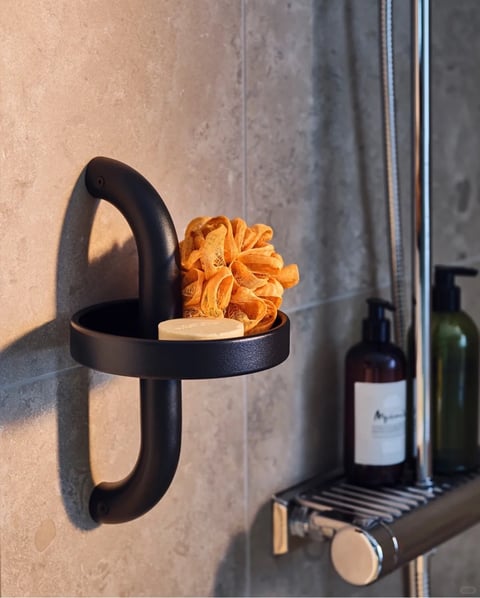 This is a photorealistic close-up shot of a modern bathroom wall, centered around a matte black, curved metal soap dish holder mounted on textured beige tiles. The holder features a thick, U-shaped bracket and a shallow circular tray, holding a pale cream-colored bar of soap and a bright orange, ruffled mesh shower pouf. To the right, slightly out of focus, sits a dark amber pump bottle with a white label and a dark green pump bottle on a chrome shower caddy. The scene is illuminated by warm, directional sunlight from the upper left, casting soft, elongated shadows and creating golden highlights on the tiles and the orange pouf, with a color palette dominated by warm neutrals, black, and vibrant orange accents. The texture of the matte black metal contrasts with the rough stone-like tiles and the soft, netted fabric of the pouf, while the composition uses a shallow depth of field to emphasize the soap holder. The only visible text is partially legible on the white label of the amber bottle, appearing as cursive script likely indicating a brand name or product description, serving to suggest a premium, natural bath product. The overall mood is calm and spa-like, with a tactile, inviting atmosphere created by the warm lighting and natural textures.
