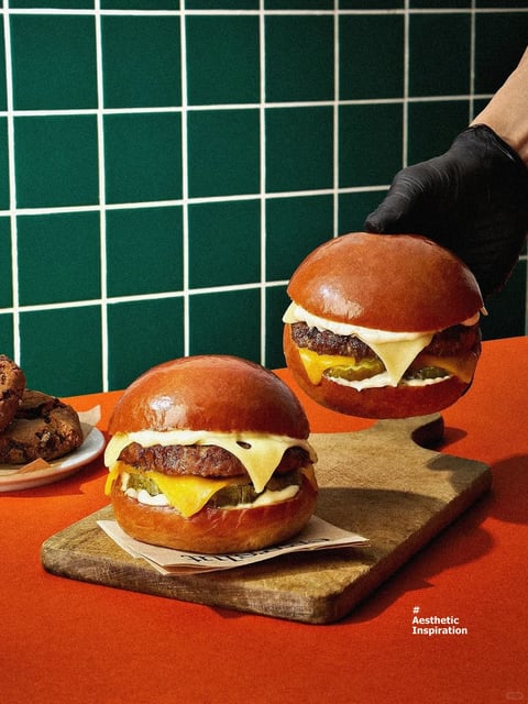 The image presents a photorealistic food photography scene with a clean, stylized aesthetic, featuring two glossy, brioche-bun cheeseburgers as the central subjects. One burger sits on a piece of branded paper atop a rustic, textured wooden serving board on the left, while a second, identical burger is held aloft by a hand wearing a black nitrile glove on the right, creating a dynamic sense of action and presentation. The composition is tightly framed against a bold, solid orange table surface and a backdrop of deep emerald green tiles arranged in a crisp white grid pattern. The color language is highly saturated and high-contrast, dominated by the warm, vibrant orange, the cool, rich green of the tiles, the deep mahogany-brown of the toasted buns, and the bright yellow of the melted cheese. Lighting is directional from the upper left, casting soft but distinct shadows that define the rounded shapes of the burgers and highlight the glossy sheen on the buns, creating a vibrant, appetizing, and clean mood. The texture rendering is highly detailed, emphasizing the smooth, lacquered surface of the buns, the crinkled, glistening surface of the meat patty, and the smooth, opaque melt of the cheese slices. Geometric shapes dominate the background with the square tiles and grid lines, contrasting with the organic, rounded forms of the burgers. In the bottom right corner, small white text reads "# Aesthetic Inspiration," indicating the image's intended use as a style or mood reference.
