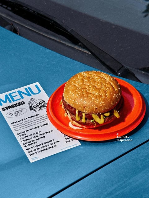 The image presents a vibrant, sunlit still life of a substantial hamburger on a bright red plastic plate, placed next to a printed menu on a blue surface, likely the hood of a vehicle given the visible windshield wiper in the background. The composition is angled from above, creating a dynamic, casual perspective. The hamburger features a glossy, sesame-seed-covered bun with a visibly textured surface, revealing layers of melted yellow cheese, a beef patty, lettuce, tomato, and possibly bacon or onion, emphasizing a hearty, indulgent appearance. The menu, positioned to the left, is a white card with bold blue and black text reading 'MENU' at the top and 'STACKED' below it, followed by a list of items such as 'STACKED AF BURGER,' 'STACKED 'N' SMOTHERED FRIES,' and 'CHICK 'N' STACK,' with prices like '$4.5' and '$15.99' visible, indicating a casual dining establishment. The visual aesthetic is photorealistic with high saturation and contrast, characteristic of a bold, retro-inspired food photography style. The color language is dominated by the warm, saturated red of the plate, the rich golden-brown of the bun, and the cool, vibrant blue of the background surface, creating a strong complementary color contrast. Lighting appears to be hard, direct sunlight, casting sharp shadows and creating bright highlights on the bun and plate, enhancing the textures and giving the scene a lively, daytime energy. The texture and material feel are pronounced, from the rough, seeded bun and glossy melted cheese to the smooth plastic of the plate and the matte paper of the menu. Compositionally, the hamburger is the central focal point, placed slightly off-center on the red plate, with the menu providing context and balance; the diagonal line of the windshield wiper adds depth and frames the scene. Shape language combines the organic, rounded forms of the burger with the geometric circles of the plate and the rectangular menu, with clean, defined edges. The mood is energetic, appetizing, and visually striking, evoking a sense of casual, indulgent dining with a nostalgic, aesthetic flair.