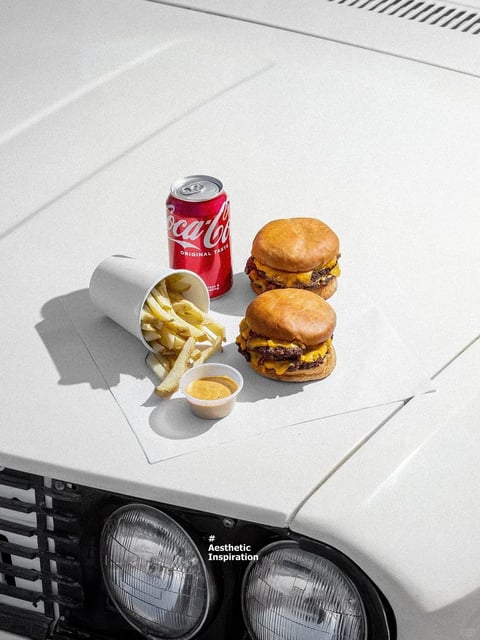 The image features a high-angle, photorealistic shot of a casual fast-food meal arranged on the flat, white hood of a vintage car, positioned just above its dual round headlights and black grille. The main subjects are two cheeseburgers with glossy buns and visible melted yellow cheese, a tipped-over white paper cup spilling golden french fries onto a white paper liner, a small plastic cup of yellow-orange dipping sauce, and a red can of Coca-Cola. Visible text includes the white cursive 'Coca-Cola' logo and the smaller text 'ORIGINAL TASTE' on the red can, which acts as a brand identifier and adds a classic commercial element. The composition is tightly framed, with the food items clustered centrally on the hood, creating a shallow depth of field where the car's headlights and grille below are slightly less focused. The visual aesthetic is clean, crisp, and highly realistic, with a bright, high-contrast color palette dominated by the white hood, the red of the can, the warm browns and yellows of the food, and the deep black of the car's grille. Lighting is hard and direct, coming from the upper left, casting sharp, well-defined shadows to the right of each object, which emphasizes the textures—the greasy sheen on the burgers, the crispness of the fries, the smooth metal of the can, and the matte finish of the paper. The mood is casual, summery, and nostalgic, evoking a sense of a quick, satisfying meal during a road trip or outdoor stop.