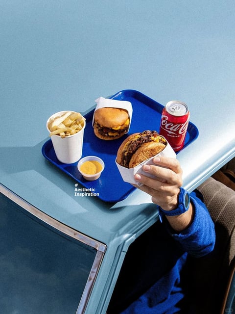 This image presents a stylized, photorealistic scene centered on a classic fast-food meal arranged on a vibrant blue tray, set against a matching pale blue tabletop. The composition is framed from a high-angle, slightly diagonal perspective, showing a person's arm and hand holding a messy, double-patty cheeseburger wrapped in white paper, positioned in the lower right foreground. On the tray, there is another similar burger in a paper holder, a white cup filled with thick-cut fries, a small plastic cup of yellow sauce, and a red Coca-Cola can. The visual aesthetic leans into a clean, graphic quality with smooth surfaces and sharp, defined edges, reminiscent of contemporary food photography or a 3D render. The color language is dominated by a cool, desaturated blue palette for the background and tray, which creates a high-contrast, complementary backdrop for the warm, saturated yellows, browns, and reds of the food and drink. Lighting is bright, diffuse, and even, casting soft, subtle shadows that suggest an overhead source, contributing to a clean, commercial mood. Texture and material feel are emphasized, with the matte finish of the tray, the glossy sheen of the soda can, the soft, porous texture of the burger buns, and the crispy, irregular surfaces of the fries all rendered with tactile precision. The composition utilizes a shallow depth of field, with the held burger in the foreground in sharp focus and the tray items slightly softer, guiding the viewer's eye. Shape language combines the organic, irregular forms of the food with the strict geometric lines of the tray and table edge. The overall mood is appetizing, nostalgic, and visually satisfying, with a high-energy contrast between the cool environment and warm food. Visible text includes the iconic white 'Coca-Cola' script logo and the word 'ORIGINAL' in smaller text on the red can, as well as a white hashtag and text overlay in the lower left reading '# Aesthetic Inspiration', which explicitly frames the image's intended style.