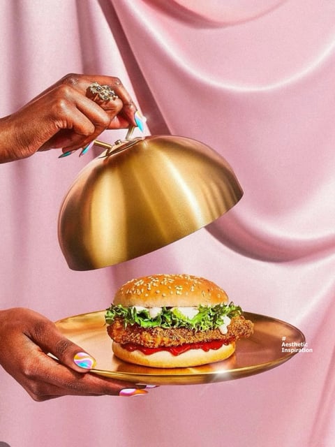 This is a stylized, photorealistic photograph featuring a pair of hands with iridescent, marbled nail polish holding a golden serving platter and cloche against a soft, draped pink fabric background. The composition is vertically oriented, with the hands positioned in the upper left and lower left, creating a diagonal flow toward the central subject: a golden-brown chicken sandwich on a sesame seed bun, garnished with lettuce, white sauce, and red sauce, resting on the reflective gold platter. The upper hand, adorned with a large, ornate ring, lifts the polished gold cloche to reveal the sandwich, creating a sense of elegant presentation. The visual aesthetic is luxurious and vibrant, with a photorealistic rendering that emphasizes glossy, reflective surfaces. The color language is dominated by a warm, monochromatic gold palette for the serving ware and sandwich, contrasted against the cool, pastel pink of the backdrop and the vivid, cool-toned iridescence of the nail polish. The lighting is soft and directional, coming from the upper left, casting gentle highlights on the gold surfaces and creating soft shadows that enhance the three-dimensional form of the hands and objects, conveying a mood of indulgence and theatrical reveal. Texture and material feel are highly emphasized, with the smooth, metallic sheen of the gold contrasting the rough, crispy texture of the chicken patty, the soft fabric folds, and the glossy nail polish. The composition uses a shallow depth of field, keeping the sandwich and hands in sharp focus while the pink background is softly blurred, directing attention to the main subject. The shape language combines the organic curves of the hands, lettuce, and draped fabric with the strong geometric hemisphere of the cloche and the circular platter. The overall mood is glamorous, playful, and aspirational, evoking a high-end, aestheticized take on fast food. In the lower right corner, there is small, white text in a sans-serif font that reads "#Aesthetic Inspiration", which serves as a descriptive tag or label for the image's style.
