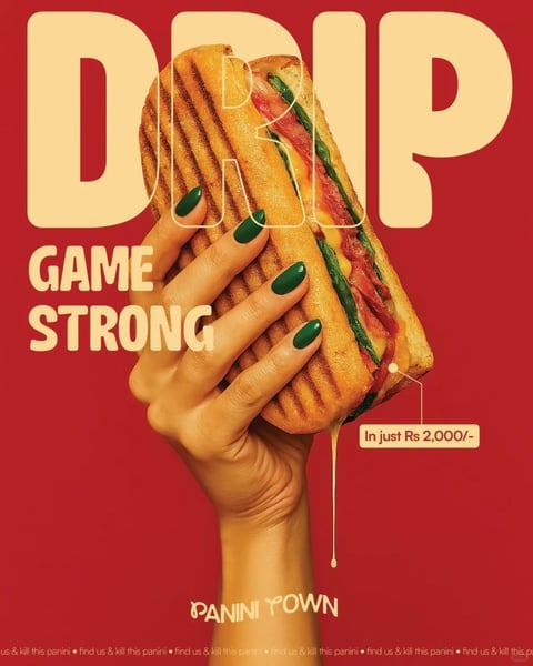 The image is a high-impact, vertically oriented advertisement featuring a photorealistic hand with glossy, dark green manicured nails gripping a large, grilled panini sandwich. The composition is centered and dramatic, with the hand emerging from the bottom edge against a solid, vibrant red background. The panini itself is a dominant visual element, showcasing deep, parallel grill marks on golden-brown bread, with visible layers of melted cheese, red sauce, and green spinach leaves, some of which appear to be dripping with a glossy, liquid cheese substance. The visual aesthetic is hyper-polished and commercial, combining realistic food photography with graphic design elements. The color language is bold and high-contrast, dominated by the saturated red background, the warm golden-browns and yellows of the sandwich, and the striking dark green of the nails, creating a vibrant and energetic palette. Lighting is directional and hard, coming from the upper left, casting strong highlights on the hand and the sandwich's surface, enhancing the textures and creating a sense of three-dimensional depth. Texture and material feel are emphasized: the sandwich shows a crispy, slightly oily bread surface with defined grill grooves, while the hand's skin appears smooth with realistic details, and the nails have a high-gloss, plastic-like finish. Compositionally, the image uses a tight, central crop focusing on the interaction between the hand and the food, with the word "DRIP" in large, bold, cream-colored sans-serif letters partially obscured behind the sandwich at the top, and the phrase "GAME STRONG" in similar typography to the left. A small text box with a leader line points to the dripping cheese, reading "In just Rs 2,000/-". At the bottom, the brand name "PANINI TOWN" appears in a stylized, slightly irregular serif font, and a repeating micro-text line along the very bottom edge reads "find us & kill this panini". The overall mood is confident, trendy, and indulgent, with a high-energy, youthful atmosphere conveyed through the bold colors, dynamic typography, and the 'drip' slang reference in both the text and the visual of melting cheese.
