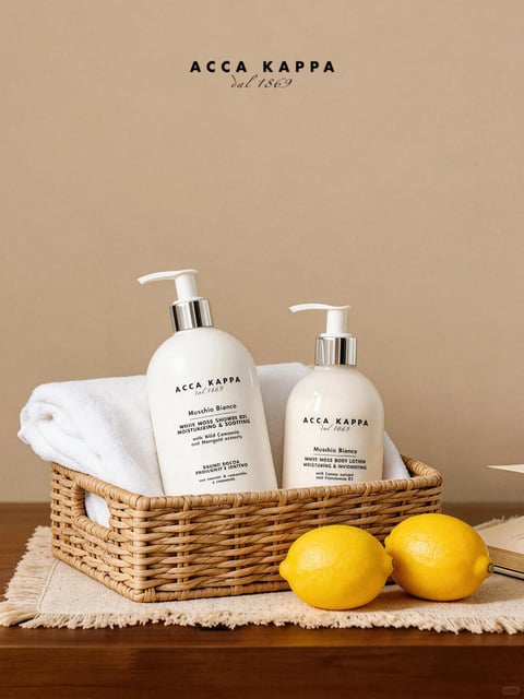 The image presents a photorealistic, styled product shot featuring two white pump bottles from the brand ACCA KAPPA, arranged within a woven wicker basket against a soft, neutral beige backdrop. The composition is centered and balanced, with the basket placed on a frayed-edge linen mat atop a dark wooden surface, accompanied by two bright yellow lemons in the foreground and a rolled white towel nestled behind the bottles. The overall visual aesthetic is clean, minimalist, and elegant, emphasizing natural textures and a serene, spa-like atmosphere. Visible text includes the brand name 'ACCA KAPPA' in a bold, sans-serif font and the tagline 'dal 1869' in a smaller, italicized script, both appearing at the top of the image and on the bottle labels; additional product text on the labels includes 'Muschio Bianco', 'WHITE MOSS SHOWER GEL', and 'WHITE MOSS BODY LOTION' in a mix of uppercase and smaller descriptive fonts, all in black ink. The lighting is soft and directional, casting gentle shadows to the right, enhancing the three-dimensional form of the bottles and the tactile quality of the woven basket, towel, and lemon rinds. The color palette is warm and muted, dominated by creamy whites, natural browns, and the vibrant pop of lemon yellow, with low saturation and high contrast between the bright objects and the darker wooden base. Textures are rendered with high fidelity, from the smooth, matte finish of the plastic bottles and the intricate weave of the basket to the plush terry cloth of the towel and the slightly porous, organic surface of the lemons. The shape language combines the cylindrical geometry of the bottles with the organic, irregular forms of the basket, lemons, and towel, creating a harmonious balance. The mood is calm, refreshing, and inviting, evoking a sense of natural luxury and self-care.