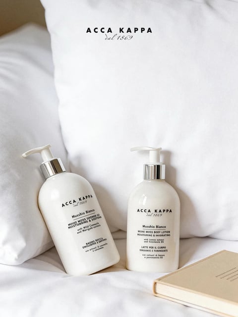 The image is a photorealistic, high-key still life photograph featuring two white pump bottles of ACCA KAPPA Muschio Bianco bath products arranged on rumpled white bedding, with a third bottle partially visible in the blurred background. The composition is intimate and centered, with the bottles placed slightly off-center on the soft, textured surface of a white pillow and sheets, creating a serene, luxurious, and domestic mood. The lighting is soft and diffused, coming from the upper left, casting gentle shadows that emphasize the round, organic forms of the bottles and the delicate folds of the fabric. The color palette is almost entirely monochromatic, dominated by varying shades of white, cream, and subtle greys, with accents of polished silver on the bottle pumps and black text. The texture is richly detailed, contrasting the smooth, matte, slightly translucent plastic of the bottles with the crisp, woven fabric of the linens and the metallic sheen of the collars. Visible text is printed in a clean, minimalist sans-serif and serif font combination: the brand name 'ACCA KAPPA dal 1869' appears prominently on the pillow and bottles, while product details like 'Muschio Bianco,' 'MOUSSE MOSS SHOWER GEL,' and 'MOUSSE MOSS BODY LOTION' are clearly legible, serving as focal points that communicate luxury and natural ingredients. The depth of field is shallow, keeping the foreground bottles in sharp focus while the background pillow and second bottle soften into a creamy blur, enhancing the intimate, tactile atmosphere of a peaceful morning or a spa-like retreat.