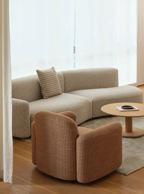 This image depicts a modern, minimalist living room scene, captured in a photorealistic style with soft, natural lighting. The composition centers on a modular, low-profile sofa upholstered in a textured, light beige bouclé fabric, featuring a single striped cushion in muted taupe and cream tones. In the foreground, a rounded armchair in a rust-orange and cream houndstooth pattern sits angled toward a simple, round wooden coffee table. The coffee table holds a small, dark bowl on top of a white book. The scene is set against a backdrop of sheer white curtains filtering bright, diffused daylight from a large window, which creates soft shadows and highlights the tactile textures of the fabrics. The color palette is warm and neutral, dominated by beige, cream, rust, and natural wood tones, with low saturation and gentle contrast. The lighting is soft and directional, coming from the window, enhancing the cozy and serene atmosphere. The shapes are predominantly organic and rounded, with smooth edges on the furniture, contributing to a gentle, inviting mood. There is no visible text or readable words in the image.