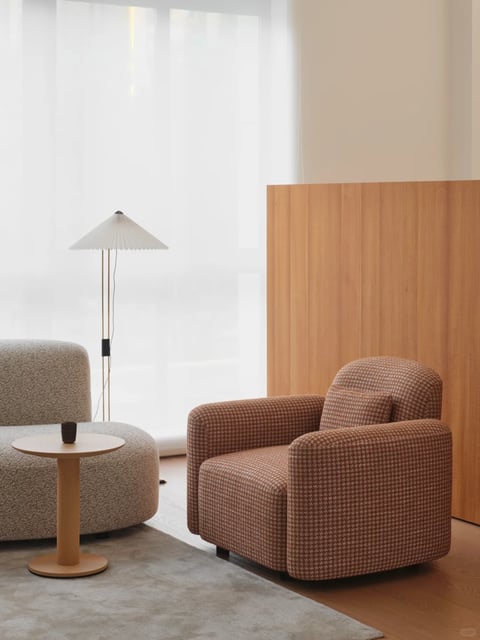 The image presents a serene, photorealistic interior scene focused on a cozy seating arrangement. The composition features a curved, textured grey sofa and a rounded armchair upholstered in a warm, brown-and-beige houndstooth fabric, positioned on a light grey rug over a wooden floor. Between them sits a minimalist, round light-wood side table holding a single dark cup. In the background, a tall, slender floor lamp with a pleated white shade stands near a large window veiled by sheer white curtains, diffusing soft, natural daylight that illuminates the scene from the left, casting gentle shadows and enhancing the tranquil mood. To the right, a vertical light-wood panel with visible grain adds a warm, geometric element. The overall aesthetic is modern and minimalist, characterized by organic, rounded shapes, a muted color palette of warm neutrals (greys, beiges, and wood tones), and a tactile interplay of textures—from the nubby sofa fabric and smooth wood to the soft curtains and pleated lamp shade. The atmosphere is calm, inviting, and balanced, with no visible text present in the image.