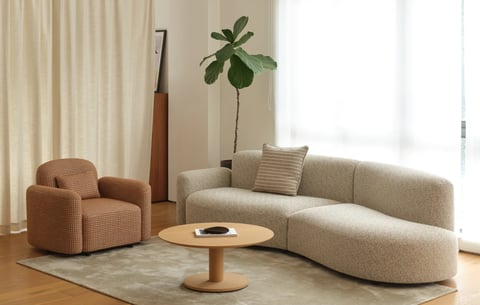 This is a photorealistic image of a modern, minimalist living room corner. The composition centers on a curved, light beige boucle sectional sofa and a matching terracotta houndstooth armchair, arranged around a light wood pedestal coffee table on a soft grey area rug. The scene is bathed in bright, diffused natural light streaming from a large window on the right, covered by sheer white curtains, creating soft shadows and a warm, serene atmosphere. The dominant color palette is a muted, warm scheme of beige, cream, terracotta, and light wood tones, with low saturation and high contrast between the bright window light and the interior elements. Textures are richly rendered, highlighting the nubby, tactile boucle fabric of the sofa, the woven texture of the armchair, the smooth grain of the wood, and the plush surface of the rug. The composition uses asymmetrical balance, with the larger sofa on the right and the smaller chair on the left, grounded by the round coffee table, creating depth through layering from the foreground rug to the background wall and window. Shapes are predominantly organic and soft, featuring rounded forms, curved lines, and pillowy silhouettes in the furniture, contrasted gently by the straight lines of the wall and window frame. There is no obvious readable text in the image, aside from a faint, indistinct graphic or logo on a magazine placed on the coffee table. A tall fiddle-leaf fig plant in the background adds a vertical element and a touch of organic green, enhancing the calm, inviting, and contemporary mood of the space.
