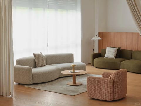 This image presents a photorealistic 3D rendering of a serene, modern living space, captured in a wide, eye-level shot that emphasizes spatial harmony and minimalist comfort. The main subject is a curated seating arrangement on a light wood floor, anchored by a soft, textured area rug. The composition is balanced, with a large, curved modular sofa in a light oatmeal bouclé fabric positioned on the left, facing a more compact, rounded armchair in a rust-and-cream houndstooth pattern to the right. Between them sits a low, round wooden coffee table with a small gold lamp and a book. In the background to the right, a secondary seating area features a deep olive-green sofa against a warm wood-paneled wall, lit by a slender floor lamp with a pleated shade. The scene is dominated by a neutral, earthy color palette of oatmeal, cream, olive green, warm wood tones, and rust, with low saturation and gentle, natural contrast, creating a soft, cohesive, and warm visual temperature. The lighting is soft and diffused, appearing to emanate from the large, sheer-curtained window on the left, casting gentle shadows and highlighting the plush, tactile textures of the bouclé, woven fabrics, and smooth wood surfaces. The shape language is predominantly organic and rounded, with soft curves on all furniture pieces, eliminating sharp edges and promoting a sense of gentle flow. The overall mood is calm, inviting, and subtly luxurious, evoking a tranquil, contemporary domestic atmosphere with low energy and high visual comfort. There is no obvious text present in the image.
