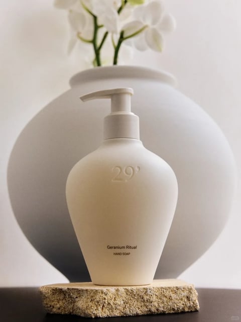 The image features a minimalist, aesthetically arranged still life centered on a beige, matte-finish pump bottle of hand soap, positioned on a rough-textured, light brown stone slab. Behind it, a large, spherical white vase with a subtle shadow and a cluster of blurred white flowers with green stems adds depth. The composition is balanced and centered, with a shallow depth of field that softens the background. Visible text on the bottle includes the embossed number "29°" near the top, and below it, the printed words "Geranium Ritual" and "HAND SOAP" in a small, clean sans-serif font, indicating the product type and scent. The visual aesthetic is photorealistic with a soft, diffused lighting approach, creating gentle highlights on the bottle's curves and soft shadows, conveying a calm, clean, and serene mood. The color language is muted and monochromatic, dominated by creamy whites, soft beiges, and earthy tones with low saturation and a warm, neutral temperature. Textures are contrasted between the smooth, matte plastic of the bottle, the porous, rough surface of the stone, and the soft, organic forms of the flowers. The shape language combines organic curves of the bottle and vase with the geometric, rough edges of the stone base, and the overall atmosphere is tranquil, elegant, and spa-like.
