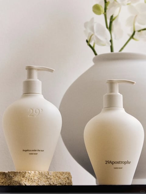 The image presents a minimalist, high-key still life composition centered on two elegant, matte-finish hand soap dispensers with pump mechanisms, set against a soft, neutral background with a large ceramic vase holding white flowers. The visual aesthetic is photorealistic with a focus on serene, modern product styling, characterized by smooth, clean lines and a tactile quality. The color language is dominated by a monochromatic palette of warm off-whites, creams, and soft beiges, with low saturation, subtle tonal contrasts, and a warm, neutral color temperature that feels organic and calming. The lighting approach is soft and diffused, appearing to come from the upper left, creating gentle gradients across the curved surfaces of the objects, minimal soft shadows, and a tranquil, airy mood. The texture and material feel are emphasized through the matte, velvety surface of the dispensers, the subtle porous texture of the stone base beneath the left bottle, and the smooth, glazed ceramic of the vase, all rendered with a clean, high-quality finish. The composition is balanced and centered, with the two bottles placed at slightly different heights on the foreground surface and the large, rounded vase filling the background, creating depth through overlapping forms and a shallow depth of field. The shape language is organic and fluid, featuring the smooth, bulbous curves of the dispensers and vase, with clean, minimalist edges and no harsh geometric lines. The overall mood and atmosphere are one of calm, sophistication, and quiet luxury, with a low-energy, peaceful visual weight. Visible text includes '29' subtly embossed on the left bottle, and 'Angelica under the sun' and 'HAND SOAP' printed in small, dark serif font near its base; the right bottle displays '29Apostrophe' and 'HAND SOAP' in a similar dark serif font, with the text serving as subtle branding and product identification within the minimalist design. The focus is uniformly sharp on the product bottles in the foreground, with the background vase and flowers softly blurred, directing attention to the soap dispensers as the primary subjects.