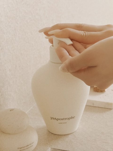 This is a soft, photorealistic close-up image centered on a white, textured ceramic pump bottle of hand soap. The scene is intimate and calming, with a person's hands positioned to press the pump, their long, manicured nails with a subtle, pearlescent finish visible. The composition is tightly framed, focusing on the interaction between the hands and the bottle, which sits on a light, neutral-toned surface against a matching, slightly textured background. The dominant color palette is monochromatic and warm, featuring creamy off-whites, soft beiges, and muted ivory tones, with low saturation and gentle contrast that creates a serene, unified feel. The lighting is soft and diffused, coming from the upper left, casting gentle shadows that define the forms without harshness and contribute to a peaceful, quiet atmosphere. Visible text on the main bottle reads "29Apostrophe" in a delicate, serif font, with "HAND SOAP" in smaller, sans-serif capitals below it; another partial bottle in the lower left shows "Angelica under the sun" and other smaller, less legible words. The overall texture is tactile and smooth yet slightly grainy, emphasizing the matte, stone-like finish of the ceramic containers and the soft, organic quality of the skin. The mood is tranquil, minimalist, and focused on a simple, everyday ritual of self-care.