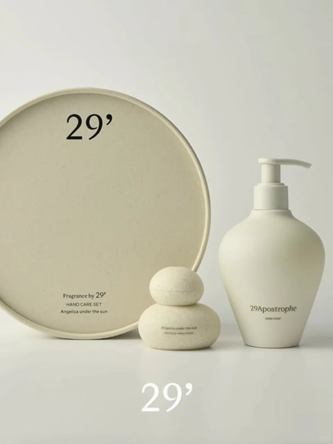 The image presents a cohesive, minimalist arrangement of personal care items, featuring three primary objects: a large, flat, round tray leaning vertically on the left, a small, stacked, pebble-shaped container at the center, and a tall, bulbous pump bottle on the right. The scene is set against a seamless, neutral off-white background with a reflective surface that subtly mirrors the base of the objects. The overall visual aesthetic is highly photorealistic and clean, evoking a sense of calm, organic luxury. The color language is monochromatic, dominated by a warm, muted beige or putty tone with a soft matte finish, exhibiting low saturation and gentle tonal contrast that emphasizes form over hue. Lighting is soft and diffused, appearing to come from the upper left, casting soft, feathered shadows to the right and below each object, creating a serene and airy atmosphere. The texture and material feel are smooth yet tactile, with a fine, speckled grain visible on the surfaces, suggesting a ceramic, stone, or matte composite material that feels both natural and refined. The composition is balanced and asymmetrical, with the large circular form on the left anchoring the arrangement, while the other two objects create a diagonal visual flow towards the right foreground, utilizing negative space effectively to convey simplicity. Shape language is predominantly organic and curved, with rounded edges and soft silhouettes on all objects, contrasting the perfect circle of the tray with the irregular, pebble-like form of the central container and the elegant, vessel-like curve of the pump bottle. The mood and atmosphere are tranquil, sophisticated, and grounded, with a low-energy, serene visual weight that feels contemplative and orderly. Visible text is integrated cleanly into the design: the large black numeral "29'" appears on the upper left of the tray; below that, in smaller black serif font, are the phrases "Fragrance by 29'", "HAND CARE SET", and "Angelica under the sun". On the central pebble container, faint text reads "Angelica under the sun" and "MEDICINAL HAND CREAM". On the pump bottle, the text "29Apostrophe" and "HAND SOAP" is printed in a similar minimalist black font. All text serves a branding and informational role, reinforcing the product identity while maintaining the minimalist aesthetic. The focus is evenly sharp across all objects, highlighting their forms and the subtle textures, with no specific area of emphasized focus, allowing the viewer to take in the entire serene composition as a unified whole.