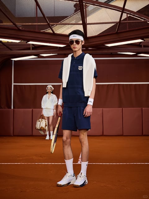 The image presents a stylized fashion scene set on an indoor clay tennis court, featuring a male model in the foreground and a female model in the background. The composition is centered on the male model, who stands facing forward with a confident, relaxed posture, holding a vintage wooden tennis racket in his right hand. He wears a navy blue polo shirt with a subtle tonal stripe pattern and a small white 'W' logo on the left chest, paired with matching navy shorts. A white knitted sweater is draped over his shoulders, secured loosely in front. His accessories include a white headband with cursive text, dark aviator sunglasses, white wristbands with script text, white crew socks, and white-and-navy tennis sneakers. The female model in the background is slightly out of focus, wearing a white short-sleeved shirt, white shorts, a white headscarf, sunglasses, and white sneakers, carrying a tan and white duffel bag. The scene is framed by the court's red-brown clay surface, a dark red padded wall, and a dark metal ceiling structure with rectangular light fixtures. Visible text includes the white 'W' logo (a stylized 'Wilson' emblem) on the polo shirt, cursive script on the white headband, and script text on the white wristbands, all in English, serving as branding elements. The visual aesthetic is photorealistic with a high-fashion, retro-sportswear mood, characterized by a warm, earthy color palette dominated by deep navy, crisp white, and the rich red-brown of the clay, with high contrast between the subjects and the background. The lighting is soft and diffused from above, creating gentle highlights on the models' skin and clothing without harsh shadows, enhancing the clean, polished feel. Textures are rendered with clarity: the smooth, technical fabric of the navy set, the soft knit of the draped sweater, the suede-like surface of the clay, and the polished leather of the sneakers. Compositionally, the image uses a shallow depth of field to emphasize the foreground model, with leading lines from the court markings and ceiling structure drawing the eye inward. Shapes are predominantly clean and geometric (the structured polo, shorts, rectangular court lines) balanced by organic forms (the models' bodies, draped sweater). The overall mood is one of sophisticated, athletic nostalgia, blending classic sportswear elegance with a modern, editorial atmosphere.