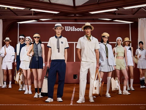 This is a photorealistic fashion editorial image set on a clay tennis court, featuring a group of eleven models styled in a sophisticated, vintage-inspired tennis aesthetic. The composition is a balanced, horizontal lineup with models arranged in two loose rows, framed by the court's net structure overhead and a dark reddish-brown backdrop. The central focus is on two male models in the foreground: one wears a cream polo with navy trim, dark trousers, and a striped tie-print scarf, holding a canvas and leather tote; the other wears a cream short-sleeve button-up, white trousers, a wide-brimmed straw hat, and a patterned neckerchief, carrying a structured leather and canvas bag. The surrounding models showcase a cohesive palette of cream, navy, pale sage green, and light blue, with outfits incorporating pleated skirts, cropped vests, blazers, and athletic silhouettes, all accessorized with visors, straw hats, pearl jewelry, and vintage-style tennis bags and rackets. The dominant color language is a warm, earthy palette centered on terracotta clay, cream, navy, and soft neutrals, with muted sage and blue accents; saturation is moderate with natural, sun-kissed tones and low contrast, creating a warm, late-afternoon glow. Lighting is directional from above and slightly front, casting soft, defined shadows that enhance the three-dimensionality of the figures and textures, conveying a calm, golden-hour ambiance. Texture and material feel are emphasized through the crisp cotton of polos and trousers, the ribbed knit of vests, the smooth leather and canvas of bags, the woven straw of hats, and the fine grit of the clay court surface. Composition is symmetrical and centered, with depth created by the layered arrangement of models from foreground to background, all anchored by the central pair. Shape language combines organic human forms with geometric lines from the clothing (pleats, stripes, structured tailoring) and the court's linear markings. The mood is polished, nostalgic, and aspirational, evoking a classic, leisurely sporting elegance. Visible text includes the word 'Wilson' in a clean, white sans-serif font centered on the backdrop, identifying the brand collaboration.