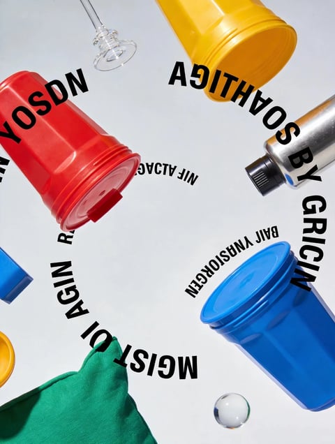 The image features a dynamic, high-angle composition of various colorful objects—red, yellow, blue, and silver cylindrical containers (some with lids), a green fabric item, and a small transparent sphere—scattered across a plain white background. The scene is arranged with an energetic, almost radial layout, creating a sense of motion or disarray. Black text in a bold, sans-serif font is overlaid in a circular pattern, with partially readable words and phrases such as "AGAIN," "RAN," "NIF ALIAC," "AGAIN," "AGAIN," "AGAIN," "BY GRCIN," "BAII YNSIOROGEN," and "AGAIN," though much appears as fragmented or nonsensical text, possibly stylized or mirrored. The visual aesthetic is clean and graphic, with a flat, illustrative rendering style that emphasizes bold shapes and solid colors. The color palette is vibrant and saturated, dominated by primary hues (red, blue, yellow) with high contrast against the white background, and a metallic silver accent. Lighting appears even and diffuse, casting soft, minimal shadows, suggesting a studio-like setup that enhances the graphic quality. Textures are smooth and plastic-like for the containers, with a matte fabric texture for the green item and a glossy, reflective surface on the transparent sphere. Compositionally, the objects and text create a circular flow, guiding the eye around the frame with strong geometric shapes and clean edges, contributing to a playful, abstract, and energetic mood with a modern, commercial art feel.