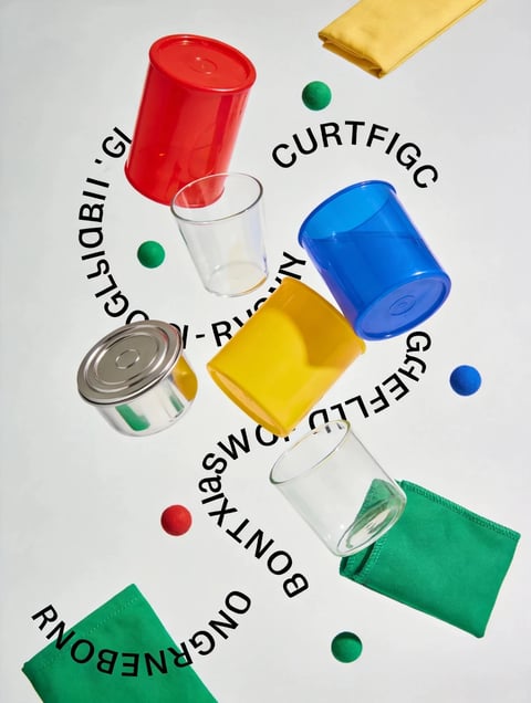 This is a high-angle, flat-lay photograph with a vibrant, graphic, and playful visual aesthetic, featuring a scattered arrangement of everyday objects against a plain off-white background. The main subjects are a collection of drinking vessels: a red plastic tumbler, a blue plastic tumbler, a yellow plastic tumbler, and two clear glass tumblers, all positioned at various angles as if frozen mid-fall. These are interspersed with a small, silver metal can, several small spherical felt balls in green, red, and blue, and folded cloth napkins in yellow and green. The composition is dynamic and circular, with the objects loosely arranged around a central, spiraling path of bold, black, sans-serif text. The visible text includes fragmented words like "CURTFIGC," "GLIABIL," "-RUS," "GFEFLD," "ONTXIS," and "ONGRNEBON," which appear to be scrambled or stylized, creating a graphic pattern that integrates with the objects rather than providing clear information. The lighting is bright, even, and soft, casting minimal shadows and creating a clean, commercial, and pop-art-inspired mood. The color language is defined by a primary palette of saturated red, blue, and yellow, accented by the green napkin and felt balls, with high contrast against the neutral background. Textures are varied but crisply rendered: the smooth, slightly glossy plastic of the tumblers, the reflective transparency of the glass, the matte metallic sheen of the can, the soft fuzz of the felt balls, and the woven fabric of the napkins. The shape language is predominantly geometric, dominated by the cylinders of the cups and can, the spheres of the balls, and the sharp rectangles of the napkins, all defined by clean, hard edges. The overall mood is energetic, abstract, and stylistically bold, emphasizing color, form, and dynamic negative space.