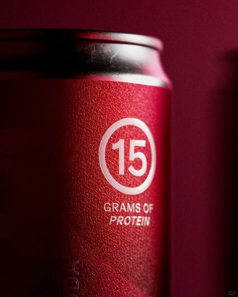 The image features a close-up, slightly low-angle shot of a red aluminum beverage can against a matching deep red background, creating a monochromatic and focused composition. The can's surface has a fine, matte, textured finish, catching light in a way that suggests a soft, diffuse illumination coming from the upper left, which highlights the can's curved shoulder and top rim while casting the right side into a gradual, soft shadow. The primary visual element is a prominent white graphic printed on the can's side: the number '15' centered within a thin white circle. Directly below this circular emblem, the text 'GRAMS OF PROTEIN' is printed in a clean, white, all-caps sans-serif font, aligned centrally. The overall aesthetic is clean, modern, and product-focused, with a high-contrast interplay between the vibrant red material and the crisp white typography. The depth of field is shallow, keeping the '15 GRAMS OF PROTEIN' text in sharp focus while the can's top and the background blur softly, directing the viewer's attention squarely to the nutritional claim.