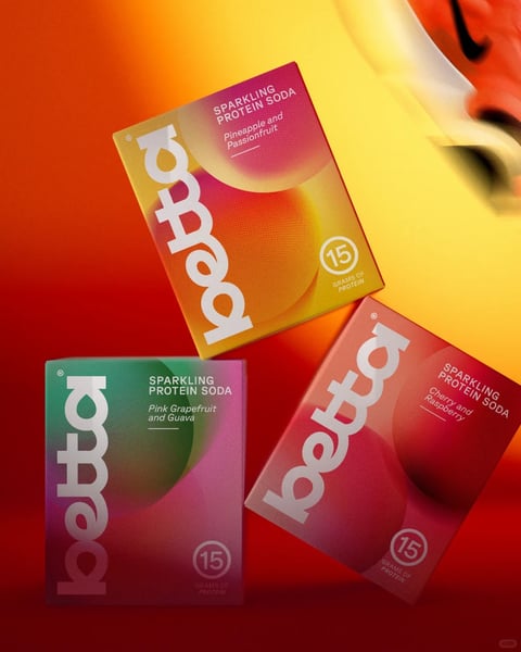 The image presents three rectangular, upright packaging boxes for 'Beota' brand sparkling protein soda, arranged in a dynamic, overlapping triangular composition against a vibrant, blurred background that transitions from deep red to bright yellow. The top box features a gradient of pink, orange, and yellow with the flavor 'Pineapple and Passionfruit', the bottom left box displays a green-to-pink gradient with 'Pink Grapefruit and Guava', and the bottom right box shows a pink-to-red gradient with 'Cherry and Raspberry'. Each box prominently features the brand name 'beota' in a bold, white, vertical sans-serif font along the left edge, and includes text such as 'SPARKLING PROTEIN SODA' and '15 GRAMS OF PROTEIN' in smaller white sans-serif fonts. The visual aesthetic is modern and graphic, with a photorealistic rendering style that emphasizes smooth, matte-finished surfaces with subtle texture and soft, curved abstract shapes in the background. The color language is highly saturated and warm, dominated by gradients of red, orange, yellow, pink, and green, creating high contrast between the vivid packaging and the warm, glowing backdrop. Lighting appears soft and directional from the upper right, casting gentle highlights on the box edges and surfaces, enhancing the three-dimensional form. The composition is asymmetrical and energetic, with the boxes tilted at various angles to create depth and movement. The shape language combines the geometric rectangles of the boxes with organic, flowing curves in the background gradients and box designs. The overall mood is vibrant, energetic, and appetizing, conveying a sense of refreshment and health-focused appeal.