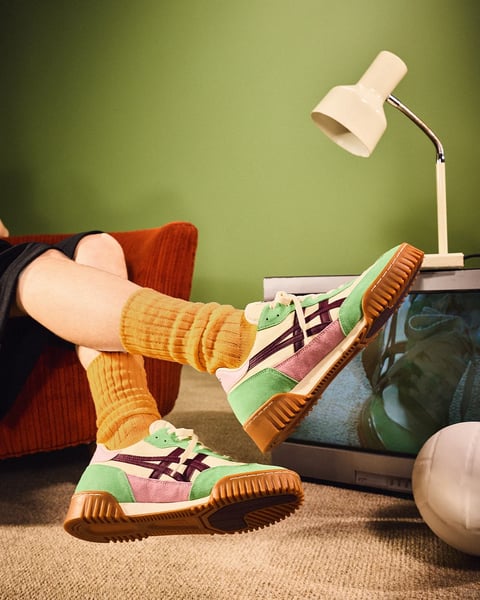The image is a photorealistic lifestyle photograph featuring a close-up, low-angle shot of a person's legs and feet, creating a relaxed, casual mood. The main subject is a pair of vibrant, retro-style athletic sneakers worn with thick, ribbed mustard-yellow socks. The person is seated in a plush, rust-orange armchair, with their legs crossed and elevated, one foot resting near a vintage television set. The scene is set in a cozy interior with a muted olive-green wall in the background and a neutral beige carpet on the floor. A white, adjustable desk lamp with a gooseneck arm is positioned on the television, casting a warm, directional light that creates soft shadows and highlights the textures of the footwear and socks. The sneakers themselves are the focal point, featuring a color-blocked design with mint green, pastel pink, and off-white panels, accented by deep purple stripes and a thick, gum-colored rubber sole with pronounced treads. The composition is tightly framed around the lower body and props, emphasizing the shoes and the interplay of colors and textures. There is no obvious readable text in the image. The visual aesthetic is warm and nostalgic, with a slightly desaturated yet rich color palette dominated by earthy tones—mustard, rust, olive green—contrasted by the brighter pastels of the shoes. The lighting is soft and warm, suggesting an indoor evening setting, enhancing the cozy atmosphere. Textures are rendered with tactile detail, from the ribbed knit of the socks and the suede-like material of the sneakers to the corduroy fabric of the chair and the matte finish of the television casing. The composition uses diagonal lines from the legs and shoes to create dynamic movement within a stable, intimate frame, while the shape language combines organic curves of the limbs and chair with the geometric lines of the shoes and television. The overall mood is comfortable, stylish, and leisurely.