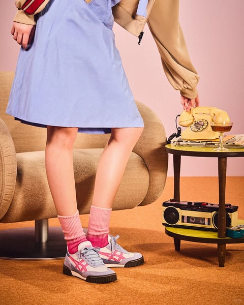The image is a photorealistic, vertically oriented studio photograph composed with a focus on a person's lower body, styled in a retro 1970s aesthetic. The subject wears a light blue, A-line dress with a flared skirt, layered under a tan bomber jacket with red and white striped cuffs. Their legs are positioned in a dynamic stance, slightly apart, with one hand reaching toward a vintage yellow rotary phone on a small, round side table. The scene is set against a soft pink backdrop and a warm, orange-brown carpet, featuring a plush, beige armchair with a curved, organic form to the left. On the side table, beside the phone, sits a cocktail glass with an amber liquid and a stack of magazines; a vintage cassette player rests on the table's lower shelf. The color language is vibrant and saturated, dominated by a warm palette of tan, orange, and pink, contrasted by the cool light blue of the dress and the white sneakers with pink accents. Lighting is soft and directional from the front-left, casting gentle shadows and highlighting the textures of the clothing and furniture. The visual aesthetic evokes a nostalgic, playful mood with a tactile feel from the plush carpet and soft fabrics. Compositionally, the frame is cropped at the subject's waist, creating a tight focus on the lower body and the interaction with the vintage objects, emphasizing shapes like the rounded furniture and the geometric lines of the table and shoes. There is no obvious readable text in the image beyond possibly indistinct markings on the magazines or cassette player labels.