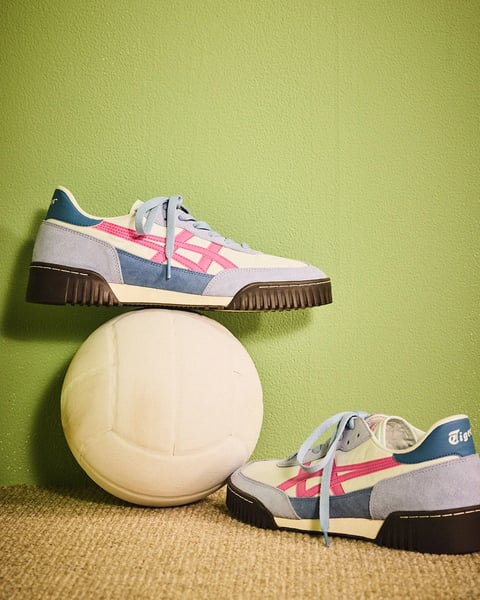 The image presents a photorealistic studio shot of a pair of retro-style sneakers, with one balanced atop an off-white, slightly worn soccer ball against a textured green wall and brown carpeted floor. The composition is asymmetrical and playful, with the upper shoe angled slightly toward the viewer and the lower shoe resting on the carpet, creating a diagonal line that guides the eye. The primary visual elements are the sneakers, featuring a color-blocked design with cream leather, light blue suede, dusty pink and teal accents, and a distinctive dark brown ribbed sole. Visible text appears on the heel tab of the lower shoe, reading 'Tiger' in a white, cursive script, indicating the brand. The lighting is soft and directional, coming from the upper left, casting gentle shadows that enhance the three-dimensional form of the shoes and ball, creating a warm, nostalgic mood. The overall aesthetic is clean and styled, with a focus on the material textures of suede, leather, and the ball's surface, rendered with high detail and a balanced color palette of muted pastels and earth tones.