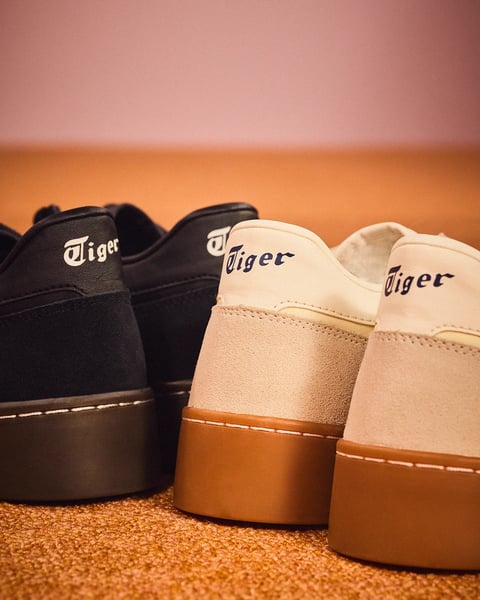 The image is a photorealistic, close-up studio shot focused on the rear heels of two pairs of low-top sneakers, arranged side-by-side on a textured, orange-brown carpeted surface. The composition is tightly framed, emphasizing the material details and branding, with the left pair in dark navy suede and the right pair in a two-tone beige suede and leather combination. Both pairs feature a distinct, stylized wordmark in a script-like font that reads 'Tiger' embroidered or printed on the heel tab, with the dark pair showing white text and the light pair displaying dark blue text. The lighting is soft and directional, coming from the upper left, casting gentle shadows and creating subtle highlights that accentuate the suede's nap and the smooth leather's sheen, contributing to a warm, nostalgic, and premium mood. The color palette is muted and earthy, dominated by deep navy, warm beige, tan, and the orange-brown of the carpet, with low saturation and high tonal contrast between the dark and light elements. The materials are rendered with clear tactile distinction, contrasting the fuzzy, matte texture of the suede panels against the smooth, matte finish of the leather heel tabs and the solid, uniform rubber of the cupsoles. The depth of field is shallow, keeping the embroidered 'Tiger' logos and heel construction in sharp focus while the background and foreground carpet blur softly, directing attention to the craftsmanship and brand identity. The shape language combines organic curves of the shoe silhouette with precise, clean stitching lines and the sharp, stylized letterforms of the branding.