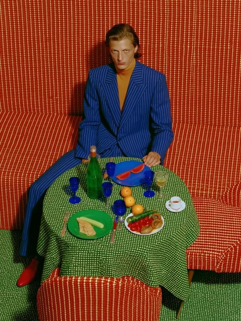 The image features a stylized, high-fashion portrait of a person seated at a small, round table, set against a visually striking, monochromatic background. The composition is centered and symmetrical, with the subject positioned directly facing the camera, framed by a vibrant red, textured wall that extends into the seating area, creating a cohesive, immersive environment. The overall aesthetic is a blend of photorealistic detail and hyper-stylized color blocking, reminiscent of pop art or a retro-futuristic editorial. The color language is bold and saturated, dominated by a primary palette of deep red, bright blue, and vivid green, with the subject's blue pinstripe suit contrasting sharply against the red background and green tablecloth. The lighting is direct and slightly harsh, casting distinct shadows and creating a high-contrast, dramatic mood that emphasizes the graphic quality of the scene. Textures are highly tactile: the red wall and seating have a fine, woven fabric pattern, the green tablecloth features a small, repeating geometric motif, and the subject's suit has a distinct pinstripe weave. Compositionally, the scene uses a flat, almost orthographic perspective with minimal depth, arranging elements like the tableware and produce in a deliberate, still-life arrangement. Shape language is a mix of geometric forms—the rectangular table, circular plates and fruits—and organic curves of the subject's posture and the food items. The mood is surreal, poised, and visually intense, evoking a sense of staged elegance with a slightly uncanny, hyper-real atmosphere. There is no obvious text or readable words present in the image.