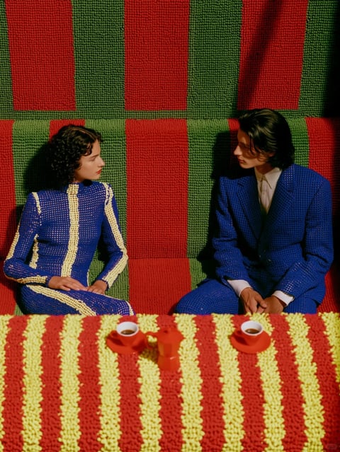 The image presents a highly stylized, photorealistic scene featuring two models—a woman with curly dark hair on the left and a man with shoulder-length dark hair on the right—seated on a textured sofa, gazing intensely at one another. They are dressed in matching blue knit suits with a fine grid pattern; the woman's outfit features bold cream vertical stripes, while the man wears a cream shirt underneath. The sofa and the table in the foreground are covered in a distinctive, tactile textile with a nubby, tufted texture resembling chenille or a deep-pile rug, featuring wide vertical stripes in vivid red and olive green on the sofa, and red and yellow on the table. On the table, two small cups of coffee sit on matching red saucers alongside a matching red pitcher, all casting distinct shadows. There is no readable text present in the image. The lighting is directional and hard, coming from the upper left, creating high contrast with deep, sharp shadows that emphasize the three-dimensional texture of the fabrics and the dramatic tension between the subjects. The composition relies heavily on geometric patterns and the repetition of vertical lines, creating a sense of order that contrasts with the intimate, charged gaze of the subjects, evoking a retro, surreal, and slightly uncanny atmosphere.