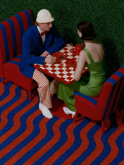 This image presents a highly stylized, photorealistic, high-angle shot of two figures seated at a small square table, evoking a retro-futuristic or pop-surrealist mood. The composition centers on a red and white checkered tabletop, flanked by a man in a vibrant blue suit with red and white striped trousers and a cream knitted hat, and a woman in a shimmering green one-shoulder dress. They sit on red chairs with vertical blue stripes, set against a dark, glittery green textured wall. The floor features a bold, wavy pattern of alternating red and blue glittery stripes. The color palette is intensely saturated and high-contrast, dominated by primary reds, blues, and greens, with sharp, hard directional lighting that casts distinct shadows, enhancing the theatrical and artificial atmosphere. The textures are rich and tactile: the glittery walls and floor, the shiny metallic fabric of the dress, the plush upholstery of the chairs, and the smooth, glossy tabletop. The shapes are a mix of clean geometric lines (the table, chair backs, stripes) and the organic curves of the figures and the wavy floor pattern. There is no visible text in the image. The overall mood is one of surreal, vibrant elegance, with a calculated and artificial energy.