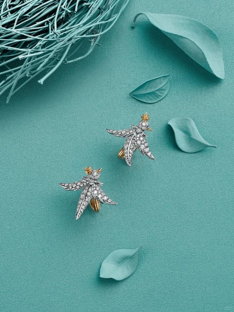 This is a flat lay photograph featuring a pair of ornate, bird-shaped jewelry pieces, likely brooches, as the central subject. The composition is arranged on a textured, teal fabric background, with additional decorative elements including a nest-like structure of painted twigs in the upper left corner and several scattered, pale green leaves of varying sizes and orientations, creating a balanced, nature-inspired scene. There is no visible text in the image. The overall visual aesthetic is highly detailed and realistic, emphasizing the intricate craftsmanship of the jewelry through a close-up, product-focused style. The color language is dominated by a monochromatic teal palette, with the background and props rendered in varying shades of cool, muted cyan and aqua. This is contrasted by the bright, metallic silver and gold of the jewelry, which is densely encrusted with small, clear, sparkling stones that catch the light. The lighting is soft, even, and diffused, appearing to come from above, which minimizes harsh shadows and creates gentle highlights on the faceted stones and metallic surfaces, enhancing the luxurious feel. The texture and material rendering are precise: the fine weave of the fabric backdrop, the matte, slightly veined surface of the leaves, the rough, painted texture of the twigs, and the brilliant, glittering texture of the pavé-set stones on the birds are all clearly discernible. The composition uses a diagonal arrangement of the two brooches, with the supporting elements framing them, creating a sense of dynamic balance and shallow depth. The shape language combines the organic, fluid curves of the birds and leaves with the geometric precision of the individual stones and the intricate, filigree-like metalwork. The mood and atmosphere are one of elegant tranquility, with a cool, sophisticated, and slightly whimsical tone conveyed through the serene color scheme and the delicate, nature-inspired subjects.