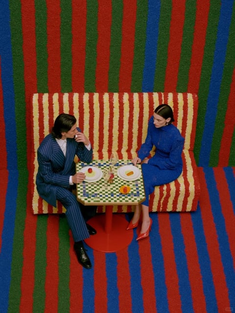 This is a high-angle, overhead shot capturing a man and woman seated across a small table, creating a stylized, theatrical dining scene. The composition is dominated by bold, vertical stripes in red, green, and blue that cover the floor and extend up the wall behind them, creating a flat, patterned backdrop. A contrasting bench with wavy, vertical stripes of cream and red runs horizontally across the middle ground. The man, on the left, wears a dark blue pinstripe suit and white shirt, holding a glass near his mouth while looking towards the woman. The woman, on the right, is dressed in a long-sleeved, high-necked royal blue dress with a textured, possibly lace-like surface, and red pointed-toe heels; she sits with hands folded in her lap, gazing downward. Between them is a small, hexagonal table with a green and white checkered mosaic top and a bright red pedestal base. On the table are two white plates, each with a small, minimalist food item—one with a red sphere on a pale base, the other with an orange segment—along with two empty wine glasses and a small, colorful decorative object. The lighting is even and bright, with minimal shadows, emphasizing the vivid, saturated colors and graphic quality of the scene. There is no visible text in the image. The overall aesthetic is highly stylized and graphic, reminiscent of a fashion editorial or art installation, with a strong focus on geometric shapes, repetitive patterns, and a bold, contrasting color palette that creates a surreal, visually striking atmosphere.